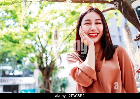 Ritratto di giovane asiatica bella donna in piedi in città con un sorriso togoloso con le mani sulla guancia guardando la macchina fotografica Foto Stock