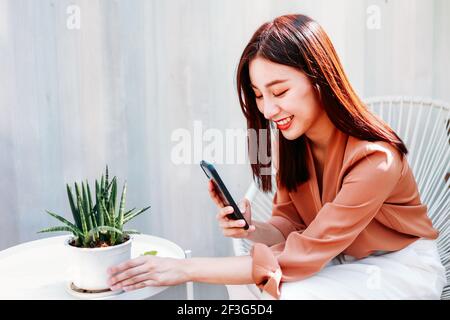 Sorridente bella giovane donna asiatica che ha un hobby di crescita nuove piante di alberi scattando foto al telefono Foto Stock