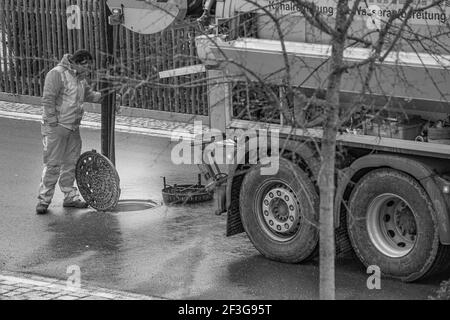 L'addetto alla pulizia delle fogne sta guardando nel tombino, in piedi dietro il suo camion con grandi ruote sulla strada in bianco e nero. Foto Stock