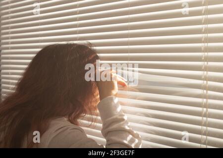 Vista posteriore di bella giovane donna con i capelli lunghi sbirciare attraverso il foro nelle tende della finestra e guarda fuori la finestra. Concetto di sorveglianza e curiosità Foto Stock