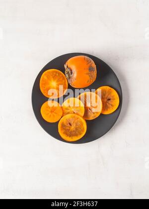 Fettine di succulenti di persimon arancio maturo e frutta intera su piatto nero su fondo testurizzato bianco. Vista dall'alto. Spazio di copia. Foto Stock