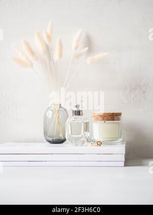 Moderna vita femminile di fiori bianchi secchi di lagurus in vaso di vetro, candela profumata di vetro e una bottiglia di profumo su una pila di riviste bianche su ligh Foto Stock