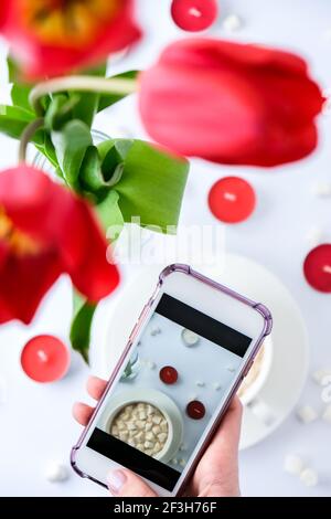Tulipani rossi candele tazza bianca di cacao marshmallows prima colazione del mattino. Mano femmina che scatta foto. Blogger. San Valentino delle madri delle donne. Vista dall'alto. Piatto Foto Stock