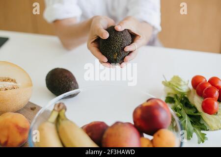 Perfetto avocado in mani sullo sfondo di frutta fresca e verdura nella moderna cucina bianca. Cucina sana e concetto di cucina casalinga. Donna che tiene Foto Stock