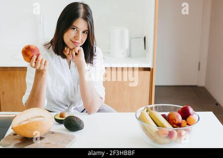 Giovane donna che tiene la pesca in mano nella moderna cucina bianca con frutta e verdura fresca sul piano di lavoro. Cucina sana e concetto di cucina casalinga. Foto Stock