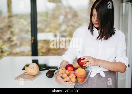 Giovane donna felice che tiene la ciotola con la frutta di estate al bancone bianco con le verdure nella cucina bianca moderna. Cucina sana e concetto di cucina casalinga Foto Stock