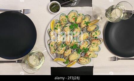 Metà delle cozze verdi Perna canaliculus della Nuova Zelanda cotte con salsa alla panna e formaggio su un piatto, decorate con spicchi di limone. Due bicchieri di whit Foto Stock