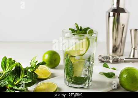 Un bicchiere di rinfrescante cocktail estivo mojito con cubetti di ghiaccio, menta fresca e lime su bianco. Ingredienti e utensili da bar per preparare mojito grink. Foto Stock