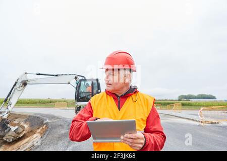 Addetto alla costruzione con computer tablet in cantiere su strada costruzione di fronte ad un escavatore Foto Stock