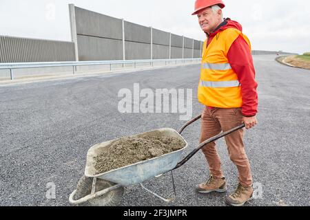 Operaio di costruzioni con carriola piena di calcestruzzo sul cantiere durante l'espansione della tangenziale Foto Stock