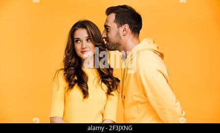 ragazzo sussurrando in orecchio di ragazza felice isolato su giallo Foto Stock