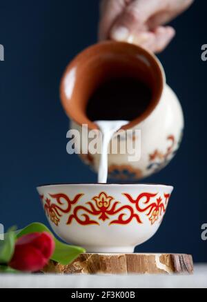 Donna kazaka che versa il latte nella tradizionale tazza da tè kese con ornamento kazako vicino leggere tulipani su sfondo blu scuro durante il festival di Nauryz. Foto Stock