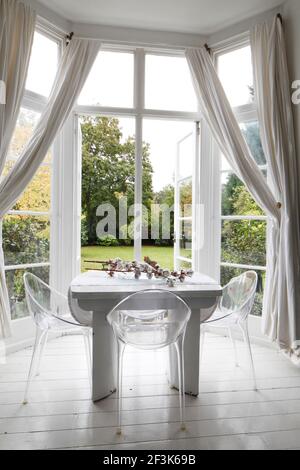 Tavolo bianco e sedie in perspex nella finestra a bovindo con drappeggi in mussola e porte aperte al giardino | | Designer: Amber Jeavons Foto Stock