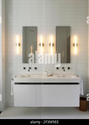 Bagno piastrellato bianco con doppio lavabo moderno, specchi e applique in casa residenziale americana. Foto Stock
