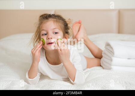 Ritratto di una ragazza sorridente felice. Cute 5 anni ragazza che si rilassa sul letto e facendo maschera facciale con fette di cetriolo. Concetto di cura della pelle. Foto Stock