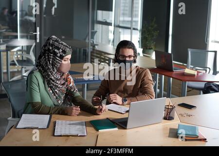 Uomini d'affari in maschere che lavorano insieme al tavolo con computer portatile in ufficio durante la pandemia Foto Stock