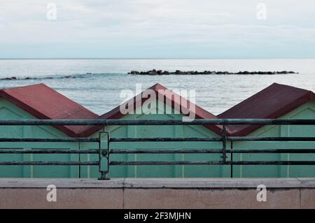 Una serie di cabine da spiaggia rosse e verdi dietro una recinzione in acciaio sulla costa mediterranea di fronte al mare (Pesaro, Italia, Europa) Foto Stock