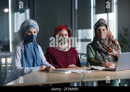 Ritratto di tre donne musulmane in abbigliamento tradizionale e protettivo maschere che guardano la fotocamera mentre lavorano insieme in ufficio Foto Stock