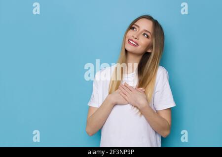 Bella giovane donna in camicia whte sta tenendo le mani sul petto, guardando via e sognando. Girata in casa su sfondo blu. Foto Stock