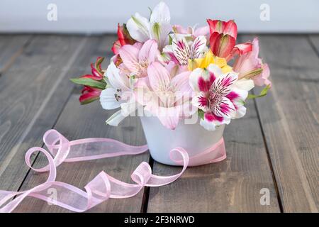 Bouquet di alstroemerie rosa, gialla, magenta, bianca e rossa in vaso bianco con nastro rosa su fondo di legno. Messa a fuoco morbida Foto Stock