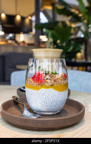 Budino di Chia con purea di mango, fiocchi d'avena e fragole fresche e tazza con cappuccino su tavolo di legno nel caffè. Messa a fuoco selettiva. Foto Stock