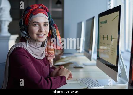 Ritratto di giovane donna musulmana in cuffia sorridente alla macchina fotografica seduto al tavolo davanti al monitor del computer Foto Stock
