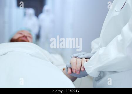 Primo piano del medico in guanti protettivi che tengono le mani del paziente e sostenendolo mentre giace su gurney Foto Stock
