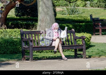 Una donna più anziana che indossa una maschera facciale leggendo il suo giornale su un banco del parco durante il blocco pandemico del coronavirus, Londra, Regno Unito Foto Stock