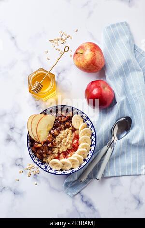 Porridge d'avena con mele caramellate con cannella, banana, fragole grattugiate e miele su sfondo di marmo chiaro, vista dall'alto. Foto Stock