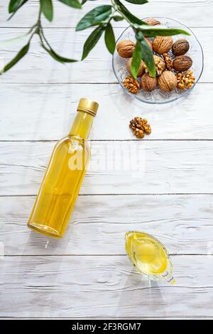 Olio di noce e un mazzo di noci su un tavolo di legno bianco. Moderna natura morta con bottiglia d'olio e recipiente in vetro con noci, rametto d'oliva e spazio per fotocopie. Foto Stock