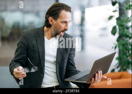 Stupito uomo adulto caucasico dai capelli grigi con computer portatile. Un uomo d'affari con un'indulgente fiducia in abiti eleganti tiene in mano un computer portatile aperto e guarda con sorpresa il monitor, togliendo gli occhiali Foto Stock