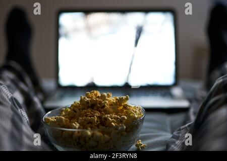 Il suo tempo di raffreddarsi. Primo piano di popcorn in un recipiente. Uomo in pigiama guardare film online con il computer portatile, sdraiato nel letto a casa la sera Foto Stock