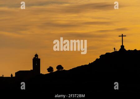 padre e figlia silhouette su una collina sotto una grande croce guardando una chiesa durante le ultime luci di crepuscolo Foto Stock