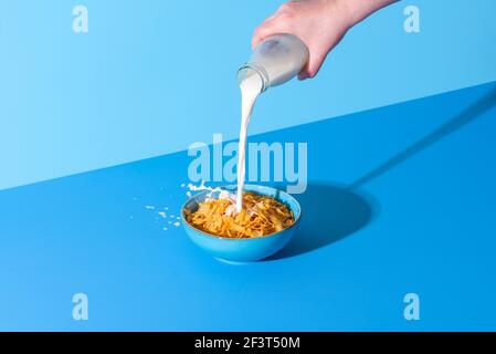 Versando il latte da una bottiglia in una ciotola con cereali a fiocchi di mais. Ciotola con cereali di mais e latte isolato su fondo blu Foto Stock