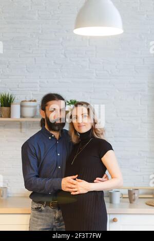 Uomo che abbraccia partner incinta in cucina. Concetto di gravidanza familiare per adulti. I futuri genitori nel vestito domestico abbracciano levandosi in piedi nella cucina, guardando e. Foto Stock