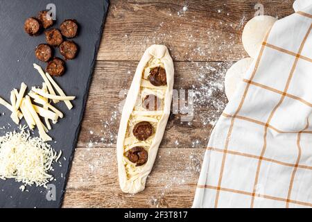 una donna sta mettendo gli ingredienti su un impasto appiattito su un piano di cucina in legno. Un'immagine concettuale per la preparazione del pane con loafs, formaggio e sucuk sul retro. Turk Foto Stock