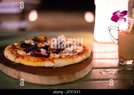 pizza con frutti di mare su un tavolo in un caffè, accanto ad un cocktail con un fiore di orchidee, una deliziosa cena sulla riva, sfondo sfocato Foto Stock