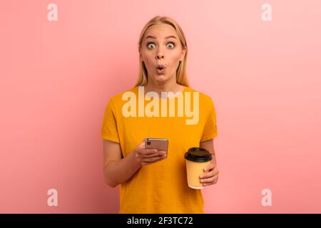 Bionda ragazza carina tipi un messaggio con il suo smartphone. Felice e sorpreso faccia di espressione. Sfondo rosa Foto Stock