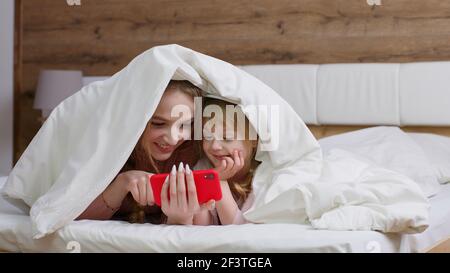 Madre sdraiata sul letto sotto il piumino con figlia ragazzina tenere il telefono cellulare guardare film online Foto Stock