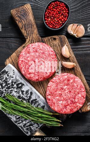 Cotolette di bistecca grezze con carne di manzo trita e rosmarino su un tagliere di legno con scagliatore di carne. Sfondo di legno nero. Vista dall'alto Foto Stock