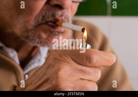 Primo piano di un uomo caucasico anziano con una barba scruffa e bicchieri neri, accendendo una sigaretta che tiene in bocca con l'accendisigari Foto Stock
