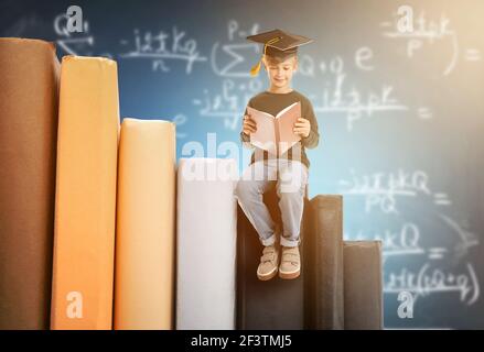 Carino ragazzo in mortaio tavola seduta su libri contro lavagna Foto Stock