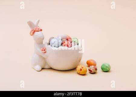 Mini caramelle al cioccolato pasquale nello stand in ceramica con coniglietto figurine su sfondo beige Foto Stock