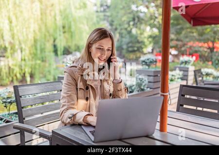 Giovane donna caucasica d'affari con capelli biondi che lavora su un computer portatile in un caffè all'aperto. Studente universitario utilizzando la tecnologia , formazione online, freelance Foto Stock