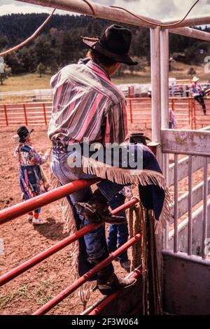 Un cowboy che gareggia nell'evento di corsa dei tori attende sulla recinzione dell'arena in un piccolo rodeo nel New Mexico. Il rodeo clown è già nell'arena. Foto Stock