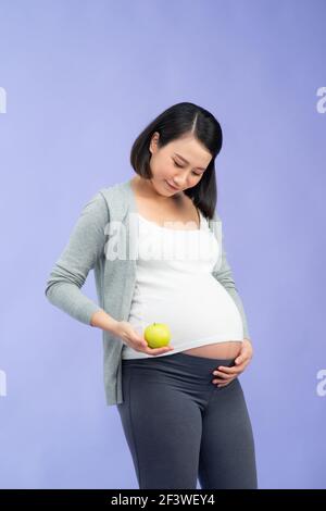 Immagine di felice giovane donna asiatica incinta in piedi isolato su sfondo viola. Fotocamera che tiene mela. Foto Stock