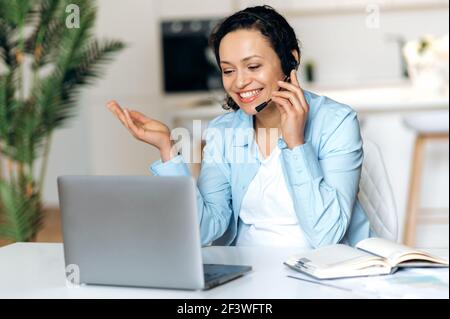 Amichevole attraente donna d'affari, freelancer o operatore di call center, in una cuffia, comunica via videochiamata con un cliente o un collega, utilizza un computer portatile mentre si siede a una scrivania, concetto di telecomunicazione Foto Stock