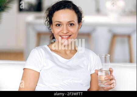 Razza mista bella donna sana in una t-shirt bianca conduce uno stile di vita sano, tiene un bicchiere di acqua pura, bevande presa d'acqua quotidiana, si siede a casa nel soggiorno, guarda la fotocamera, sorridendo Foto Stock