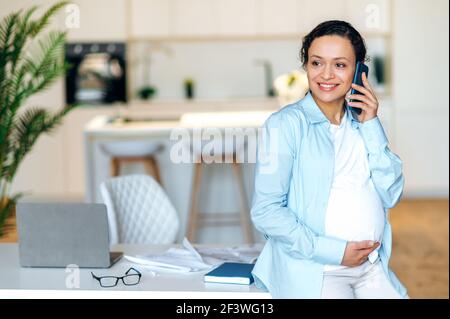 Bella razza mista donna incinta adulto è in piedi vicino al suo posto di lavoro, avendo piacevole conversazione cellulare, delicatamente toccando il suo ventre, guardando via e sorridendo Foto Stock