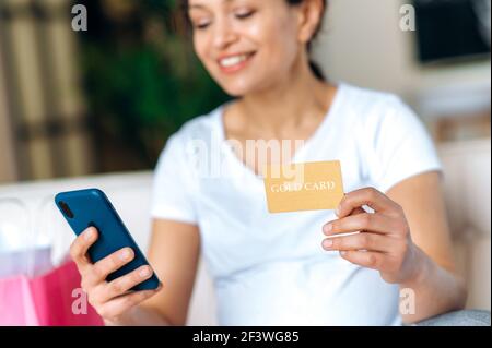 Donna incinta sorridente sfocata in una t-shirt bianca sedersi sul divano, tiene una carta di credito e uno smartphone in mano, effettua acquisti online, acquista vestiti e giocattoli per il futuro bambino, shopping online Foto Stock